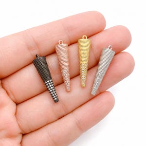 Puede incluir: Cuatro dijes de joyería en forma de cono con una superficie texturizada y piedras transparentes incrustadas. Los dijes se presentan en cuatro acabados metálicos distintos: negro mate, oro rosa, oro amarillo y plata, cada uno con una pequeña argolla.