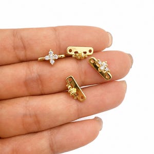 May include: Four gold-toned jewelry connectors featuring a rectangular base with two small holes at each end and a central decorative element adorned with clear, sparkling cubic zirconia stones arranged in a four-petaled flower shape.