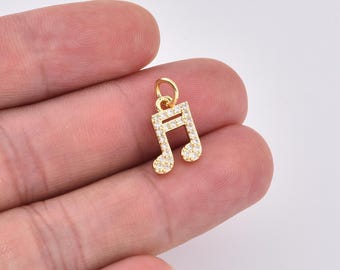 Pode incluir: Um pingente de nota musical dourada, incrustado com pequenas pedras preciosas transparentes. O pingente é uma clave de sol, com uma argola no topo para prender a um colar ou pulseira. O pingente é segurado em uma mão.