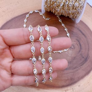 May include: Gold-toned chain with oval-shaped clear crystals. The chain is displayed in a hand, with a roll of the same chain visible in the background. The crystals are set in a bezel setting, and the chain is on a wooden surface.