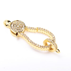 May include: Gold-tone clasp with a round, clear crystal-encrusted design. The clasp is shaped like a teardrop with a smaller, round loop at the bottom.