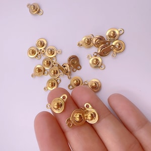 May include: A collection of small, gold-colored metal clasps. Each clasp features a rounded, button-like center and a small loop for attachment. The clasps are scattered across a light-colored surface, with some held in a hand.