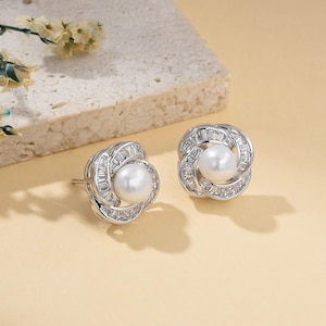 May include: A pair of silver-toned stud earrings. Each earring features a central white pearl surrounded by a flower-like design of baguette-cut clear crystals. The earrings are set against a neutral background.