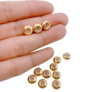 May include: A selection of gold-coloured, round beads with a hole through the centre. The beads have a smooth, shiny finish and are scattered on a white background. These beads are suitable for jewellery making and crafting.