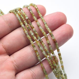 May include: Strands of small, square-cut beads in shades of olive green and brown. The beads are strung together, creating a delicate and textured appearance. The beads are held in a hand against a white background.
