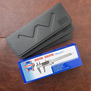 May include: Three black rectangular plastic storage cases stacked together, positioned above a blue and white cardboard box labelled DIGITAL CALIPER 150mm (6"). The box features an illustration of a stainless steel digital caliper tool.