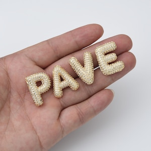 Może przedstawiać: Złote zawieszki w kształcie liter, tworzące słowo "PAVE" z przezroczystymi kamieniami cyrkonowymi.