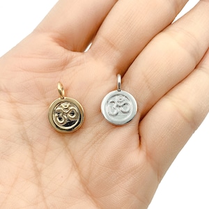 May include: Two silver charms with an Om symbol, one gold and one silver. The charms are on a hand.