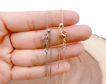 May include: Two delicate chains, one silver and one gold, with adjustable clasps. The chains are held in a hand against a wooden background.