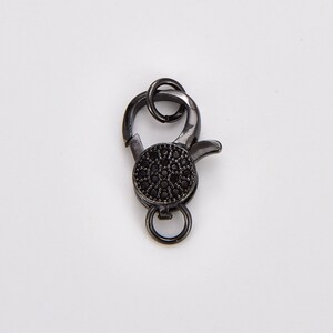 May include: Black metal clasp with a round, black, pave-set stone. The clasp has a spring-loaded opening and a loop for attaching to a chain or cord.