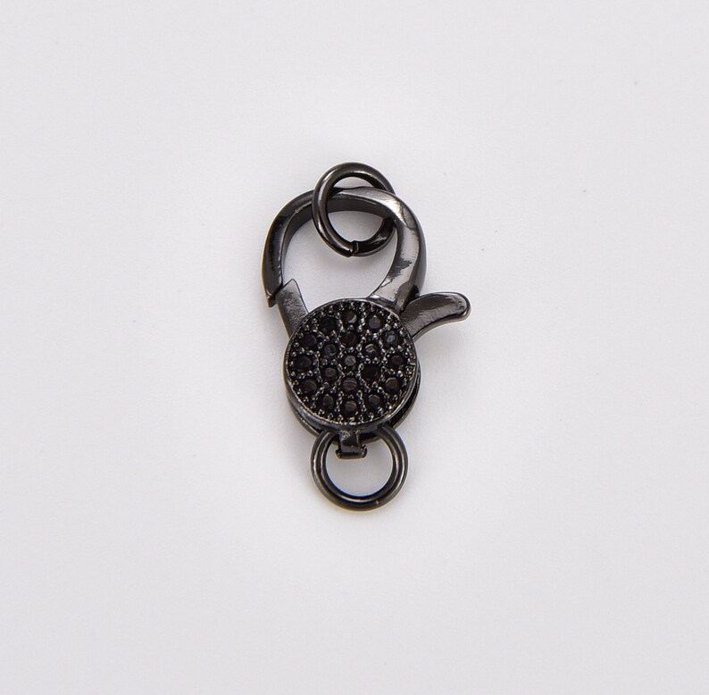 May include: Black metal clasp with a round, black, pave-set stone. The clasp has a spring-loaded opening and a loop for attaching to a chain or cord.