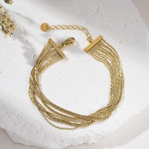 May include: A gold-colored bracelet with multiple strands of delicate chain. The bracelet features a rectangular clasp and an adjustable chain for a custom fit. The jewelry is displayed on a white, textured surface.