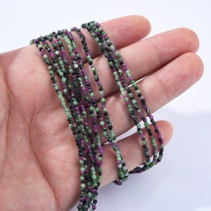 May include: Strands of faceted gemstone beads in shades of green, black, and purple. The beads are strung together, creating a delicate and colorful necklace or bracelet material. The gemstones have a polished, reflective surface.