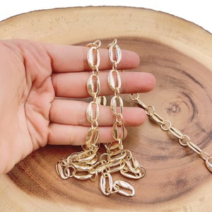 May include: A gold-toned chain necklace made of linked seashell-shaped charms. The chain is shown on a wooden surface.