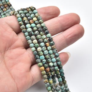 May include: Strands of turquoise gemstone beads with a faceted, square shape. The beads display a mix of green, blue, and brown hues. The beads are held in a hand against a white background. These beads are suitable for jewelry making.