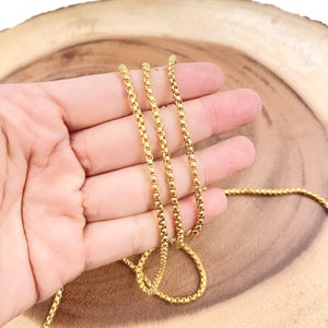 May include: A gold chain necklace with a box chain design. The chain is shown on a wooden surface.