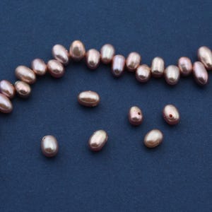Puede incluir: Perlas rosas de forma ovalada, algunas ensartadas, otras sueltas, sobre un fondo azul oscuro. Las perlas tienen un brillo nacarado y varían en tamaño. La imagen muestra la belleza natural de las perlas.