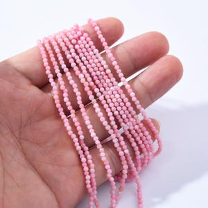 May include: Strands of small, light pink beads are held in a hand. The beads are uniform in size and shape, strung together to create a delicate, textured appearance. The beads are a soft, pastel pink color.