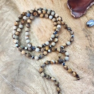 May include: A long, multi-strand necklace made of brown, white, and gray gemstone beads. The necklace is knotted with brown cord and has a silver metal accent. The necklace is lying on a wooden surface.