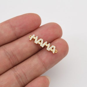 May include: A gold-colored charm spelling "MAMA" in a cursive font, encrusted with small, clear gemstones. The charm has small loops on each end for attachment. The background is a light, neutral color.