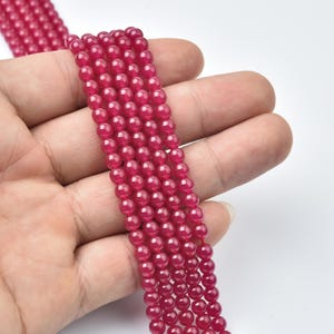 May include: Strands of small, round, ruby-red beads. The beads are smooth and evenly sized, with a glossy finish. The beads are held in a hand against a white background. The beads are strung together, ready for jewelry making.