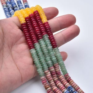 May include: Strands of colorful gemstone beads in yellow, red, green, pink, and blue hues. The beads are small, round, and strung together, creating a textured and vibrant appearance. The beads are held in a hand against a white background.