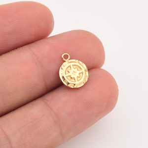 May include: A small, gold-colored charm with a compass rose design. The charm is round with a textured border and a small loop at the top for attaching to a necklace or bracelet. The compass rose is a stylized, eight-pointed star.