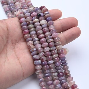 May include: Strands of faceted gemstone beads in shades of pink, purple, and white. The beads are small and round, with a polished, reflective surface. The beads are held in a hand, showcasing their color variations and texture. Ideal for jewelry making.