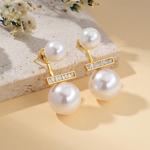 May include: A pair of gold-toned earrings featuring a large, lustrous white pearl at the bottom and a smaller pearl at the top. The earrings have a rectangular bar with small, clear stones. The earrings are set against a neutral background.