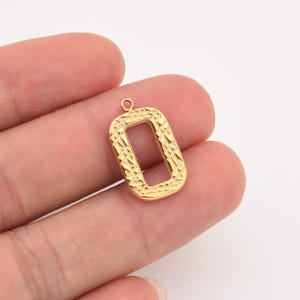 May include: A gold-colored rectangular charm with a textured surface and a small loop at the top. The charm has an open center and is held in a person's hand.