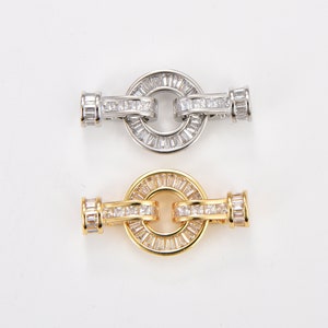 Könnte beinhalten: Zwei silberne und goldfarbene Metallverschlüsse mit klaren Kristall-Akzenten. Die Verschlüsse sind rund mit einem Kreisdesign in der Mitte.