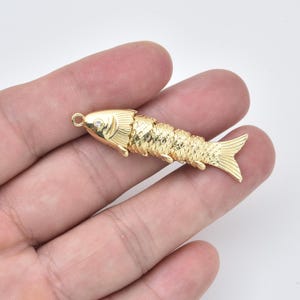 May include: A gold-colored fish pendant with a textured scale design. The fish has a small loop at the top for attaching to a chain or other jewelry. A small, clear gem is set in the fish's eye. The pendant is approximately 2 inches long.