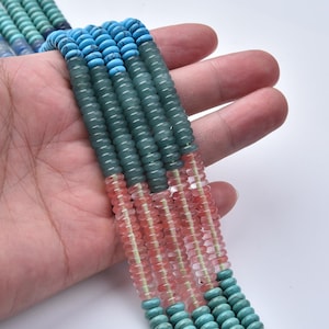 May include: A close-up of strands of colorful beads. The beads are in shades of turquoise, teal, pink, and clear. The beads are round and strung together, creating a textured and vibrant appearance. The beads are held in a hand.