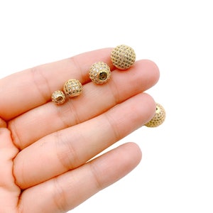 May include: Three gold-toned round beads with clear rhinestones. The beads are different sizes and are arranged in a row on a hand.
