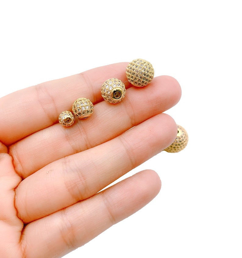 May include: Three gold-toned round beads with clear rhinestones. The beads are different sizes and are arranged in a row on a hand.