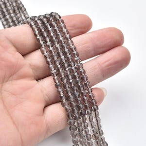 May include: Strands of faceted, smoky quartz beads. The beads are a translucent, brownish-gray color and are held in a person's hand. The beads are small and appear to be strung together, suitable for jewelry making.