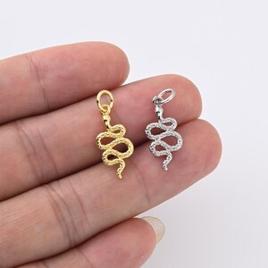 May include: Two snake-shaped charms are shown, one gold and one silver. The charms have a coiled design and a small loop at the top for attachment. The background is a light, neutral colour.