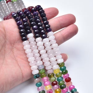 May include: A collection of gemstone bead strands in various colors, including purple, black, white, pink, green, and red. The beads are faceted and strung together, creating a colorful and textured appearance. The strands are held in a hand.