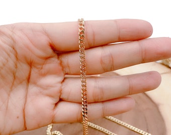 May include: A gold chain necklace with a curb link style. The chain is shown laying on a hand and a wooden surface.