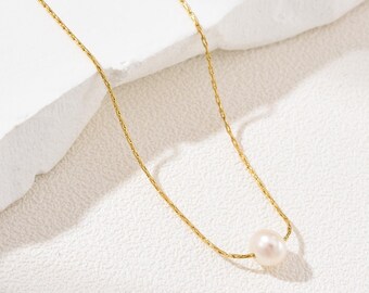 May include: A delicate gold necklace featuring a single, lustrous pearl pendant. The chain has a fine, textured design, and the pearl is a soft, creamy white. The necklace is displayed against a white, textured backdrop.