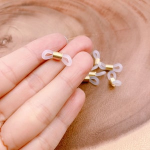 以下が含まれることがあります： 白いプラスチックと金色の金属製のリングが付いた、眼鏡チェーンコネクターを手に持っている手のアップ写真。