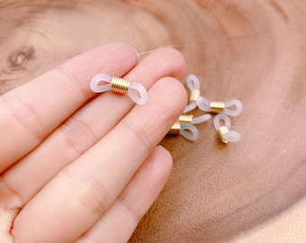 Puede incluir: Primer plano de una mano sosteniendo varios conectores de cadena de gafas blancos y dorados. Los conectores están hechos de plástico transparente con un anillo de metal dorado en el centro.