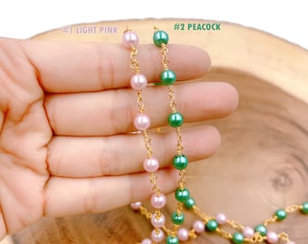 Puede incluir: Dos hebras de cadena dorada con cuentas de perlas. Una hebra tiene cuentas de color rosa claro y la otra tiene cuentas de color verde pavo real. Las hebras se sostienen en una mano.