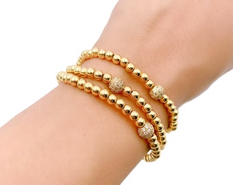 May include: Three gold beaded bracelets with small, sparkly, round accents. The bracelets are stacked on a person's wrist.