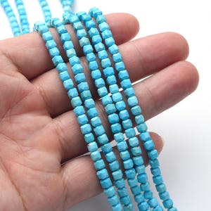 May include: Strands of light blue, faceted, square-shaped beads. The beads are strung together, creating multiple parallel lines. The beads have a slightly irregular surface, and the color is a vibrant turquoise. The beads are held in a hand against a white background.