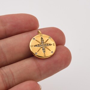 May include: A gold-colored compass pendant with a starburst design in the center, encrusted with small, clear gemstones. The compass points are marked with N, E, S, and W. The pendant is round and has a small loop at the top for a chain.
