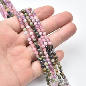 Puede incluir: Primer plano de hebras de pequeñas cuentas de piedras preciosas facetadas. Las cuentas son de color rosa, verde y negro. Las cuentas se sostienen en una mano, mostrando sus superficies pulidas y reflectantes.