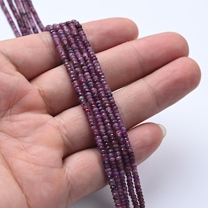 May include: Close-up of multiple strands of small, faceted, deep magenta-colored gemstone beads. The beads are held in a hand, showcasing their color and texture. The beads are strung together, ready for jewelry making.