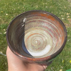 Puede incluir: Un cuenco de cerámica marrón con un patrón de remolino blanco y naranja en el interior. El cuenco está esmaltado y tiene un acabado liso.