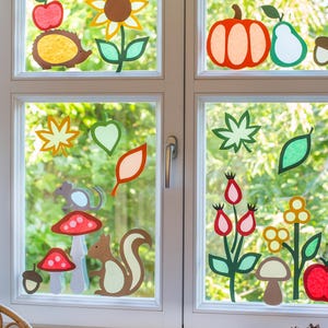 Könnte beinhalten: Ein Fenster, das mit farbenfrohen Herbstmotiven verziert ist. Die Dekorationen umfassen einen roten Apfel, eine Sonnenblume, einen Kürbis, eine Birne, Blätter, Pilze, ein Eichhörnchen und andere herbstliche Elemente. Das Fenster ist weiß gerahmt, mit einem karierten Volant oben.